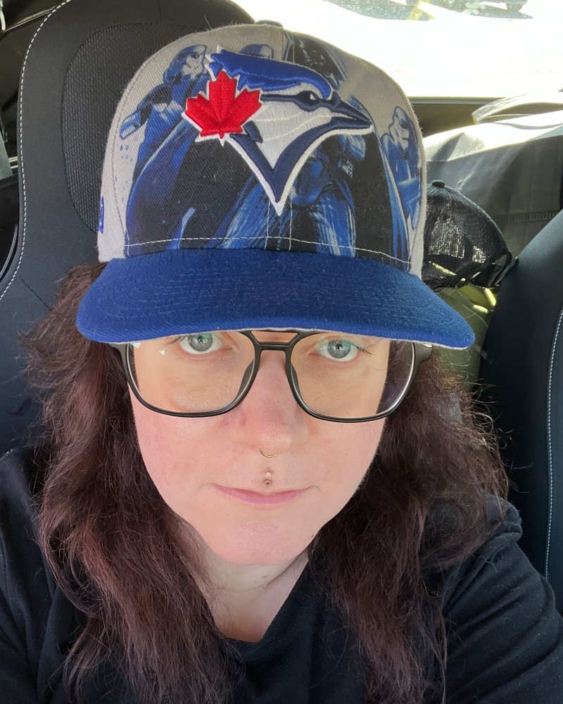 Sparrow Panton: a white person with long dark red hair, black-framed glasses, a nose ring and a lip piercing, wearing a Toronto Blue Jays cap and a black t-shirt, looking straight at the camera.